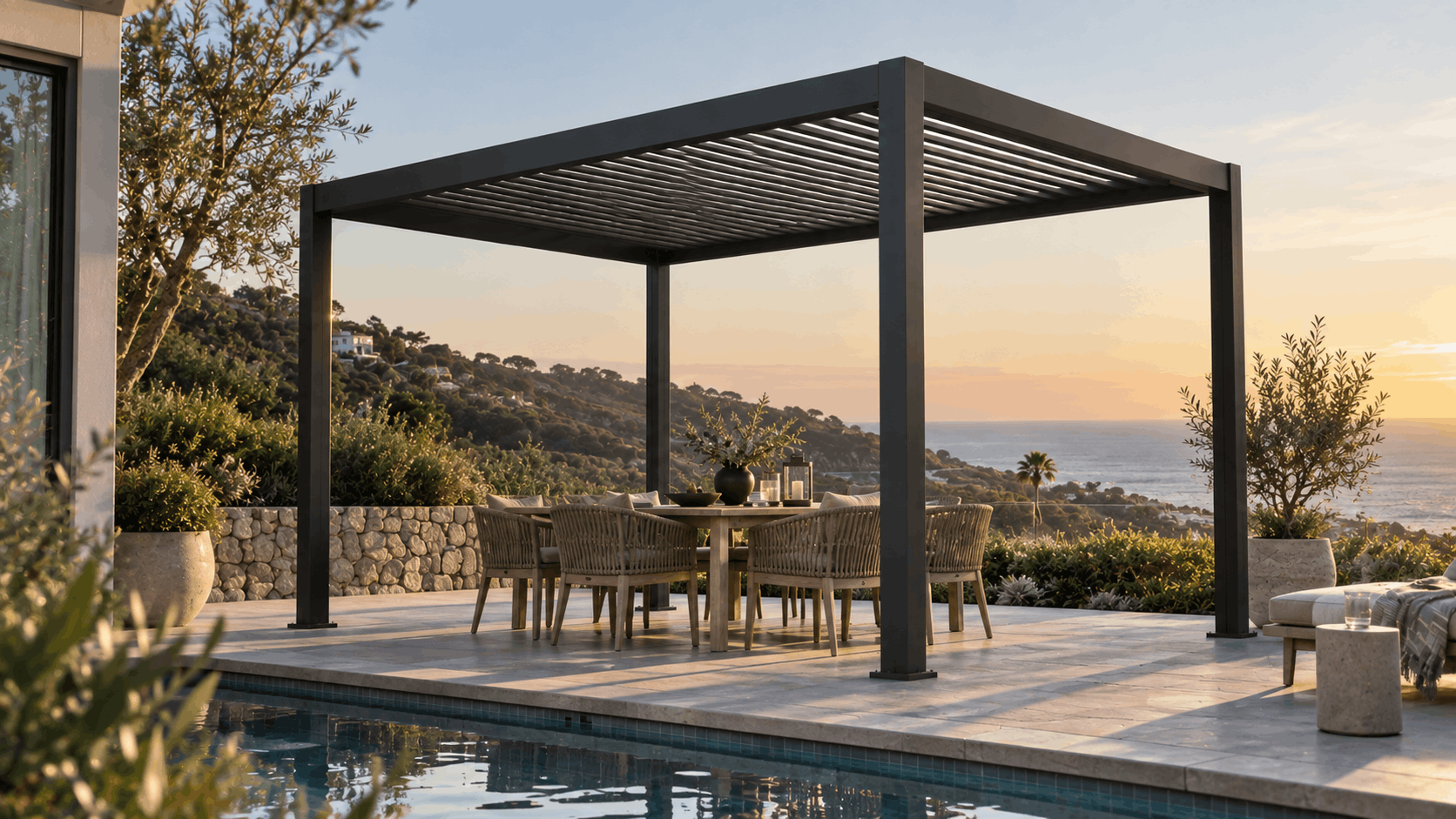 Black aluminum louvered pergola over a dining table beside a pool overlooking the ocean at sunset