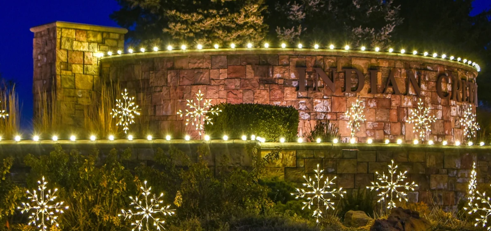 Stone community entrance sign lit with warm white string lights and illuminated snowflake displays at dusk