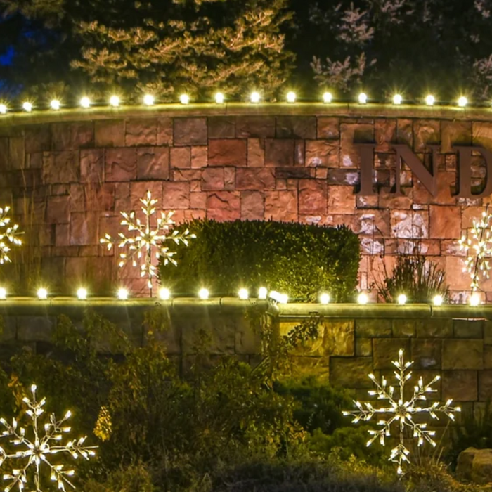 Stone community entrance sign lit with warm white string lights and illuminated snowflake displays at dusk