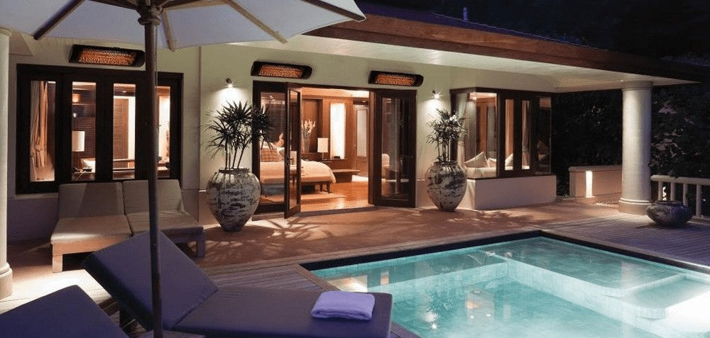 Three wall-mounted electric infrared patio heaters glowing over a covered poolside patio at a home in the evening