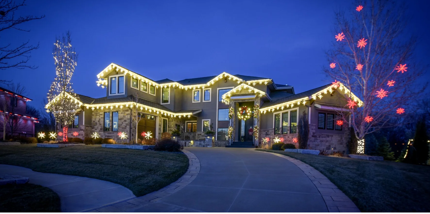 Two-story house decorated with warm white roofline Christmas lights, lit snowflake yard displays, and a holiday wreath at dusk