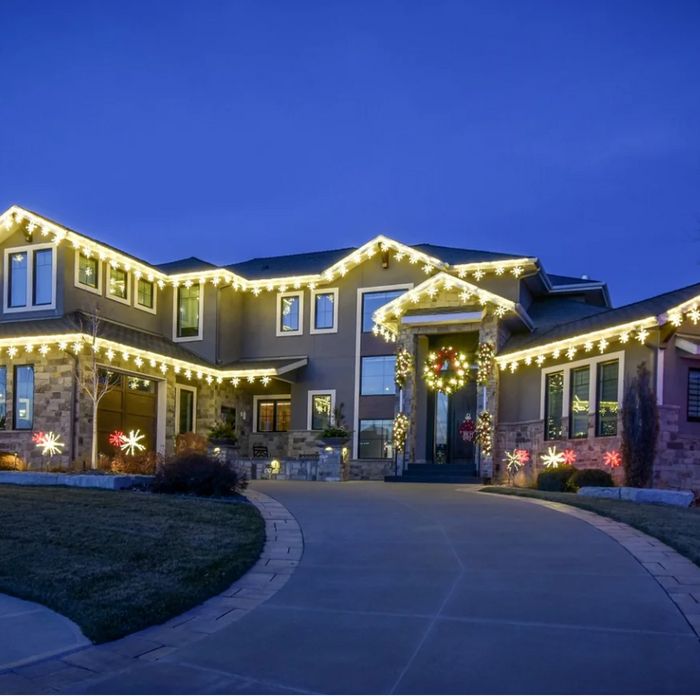 Two-story house decorated with warm white roofline Christmas lights, lit snowflake yard displays, and a holiday wreath at dusk