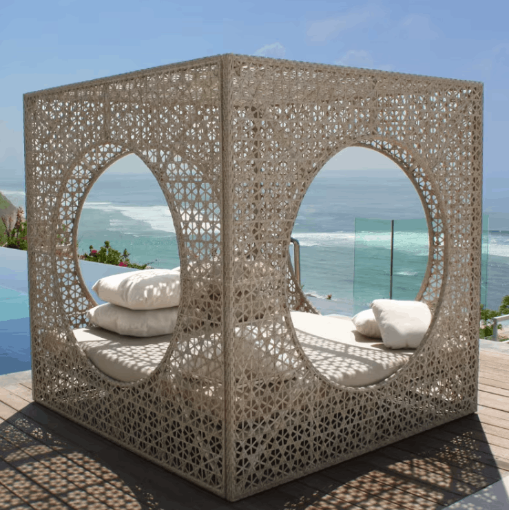 A large outdoor lounge cabana daybed with intricate geometric laser-cut metalwork, featuring two large circular openings, cream cushions, and a beachfront ocean view background.