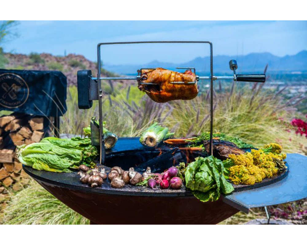 Arteflame Rotisserie Kit roasting whole chicken on spit over Arteflame Grill with fresh vegetables in outdoor desert setting