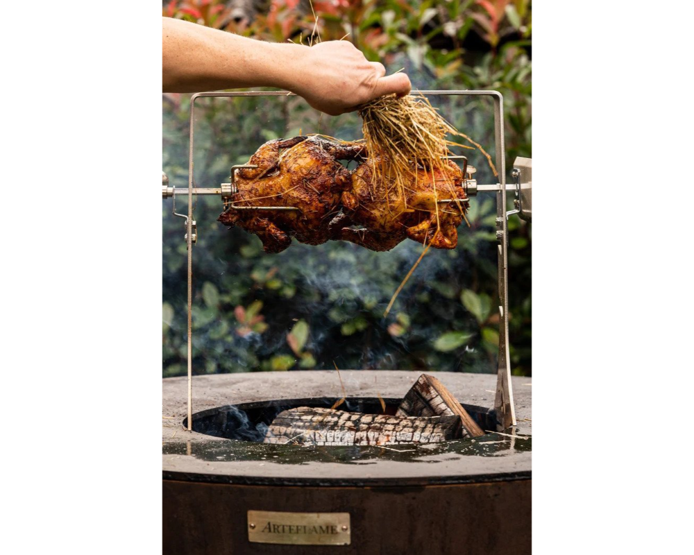 Arteflame Rotisserie Kit with golden rotisserie chicken on spit over open wood fire in Arteflame Corten Steel Grill