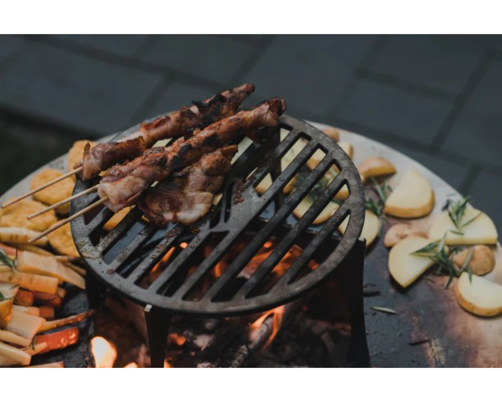 Arteflame Classic 20" Corten Steel Grill cooking meat skewers and potatoes on cast iron grate over open wood fire