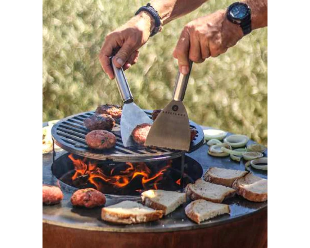 Arteflame Classic 30" Grill with person cooking burgers and bread on flat-top corten steel surface over open flame
