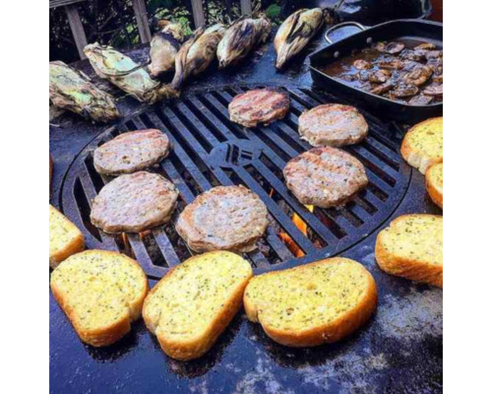 Arteflame Classic 30" Grill cooking burger patties, garlic bread, corn, and mushrooms on flat-top steel surface over fire