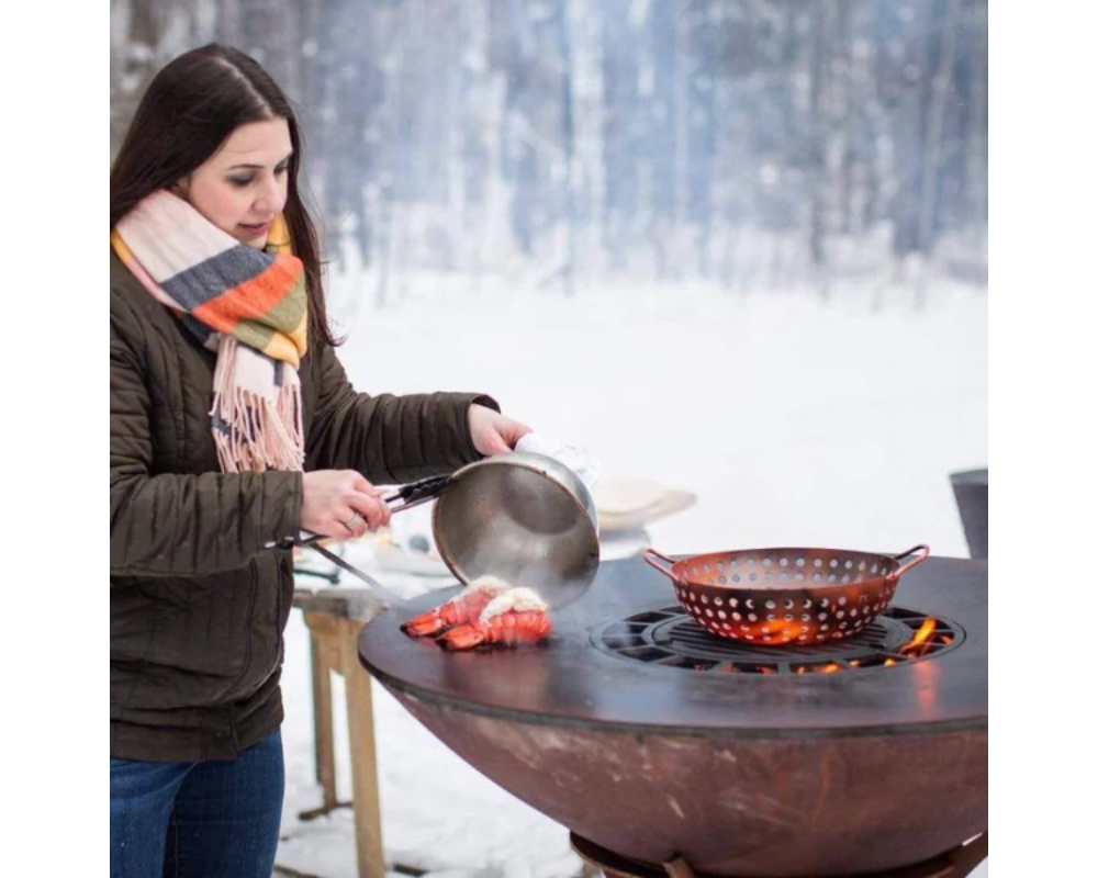 Arteflame Classic 40" Grill with woman cooking lobster tails on flat-top steel surface outdoors in winter snow setting