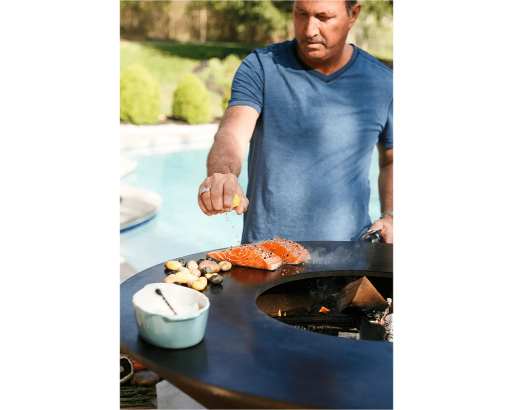 Arteflame Classic 40" Grill with man squeezing lemon over salmon and potatoes on flat-top steel surface by pool