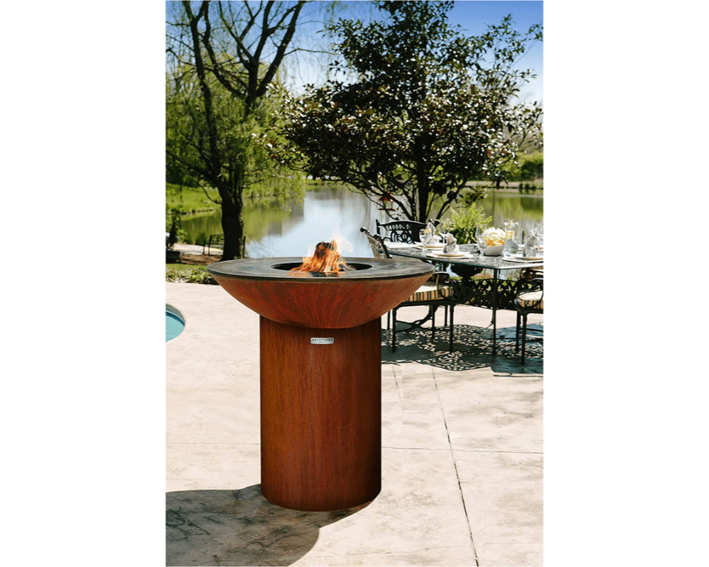 Arteflame Classic 40" Corten Steel Grill with Tall Round Base on outdoor patio by pool and lakeside dining setting