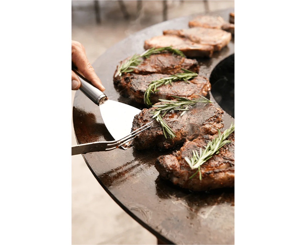 Arteflame Classic 40" Grill flat-top steel surface searing ribeye steaks with fresh rosemary and stainless steel spatula