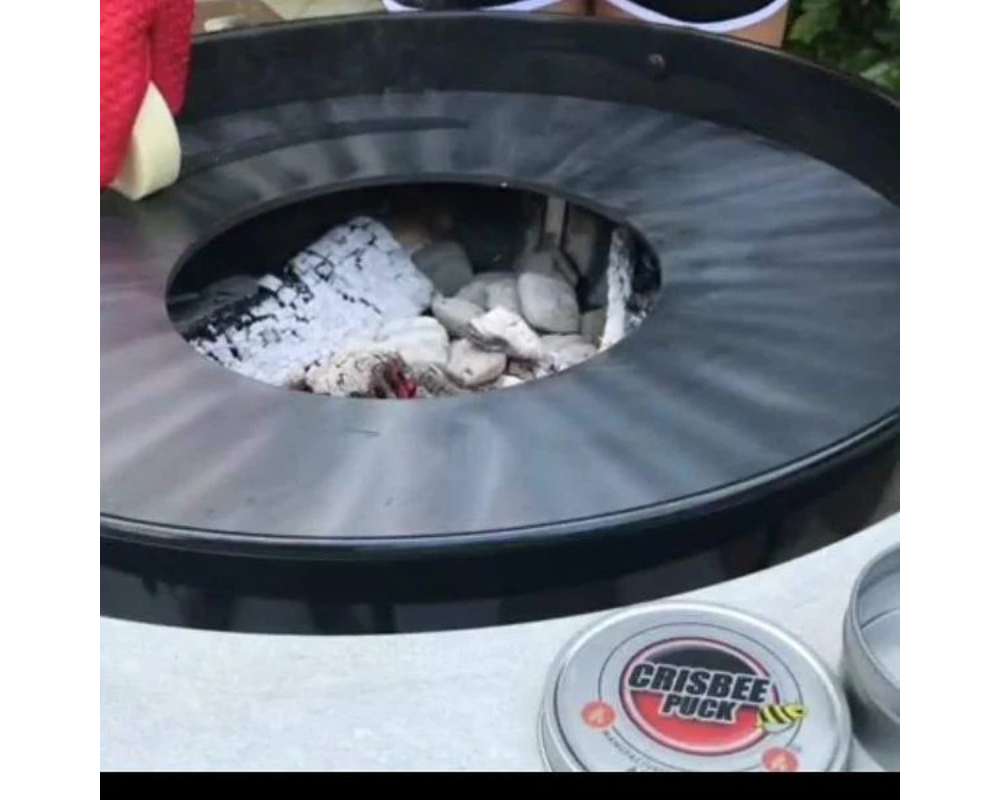 Crisbee Puck grill seasoning tin beside Arteflame carbon steel flat-top cooktop with ash-filled center fire bowl