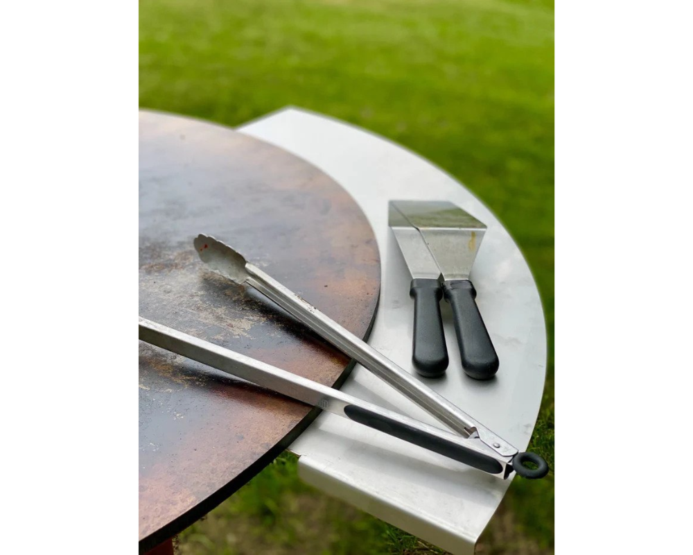 Arteflame Stainless Steel Side Warming Table shelf holding grill tongs and spatula beside Arteflame corten steel cooktop