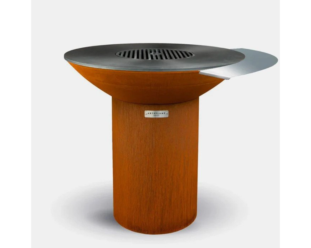 Arteflame round outdoor grill with dark steel cooktop, side shelf, center grate, and cylindrical corten steel base