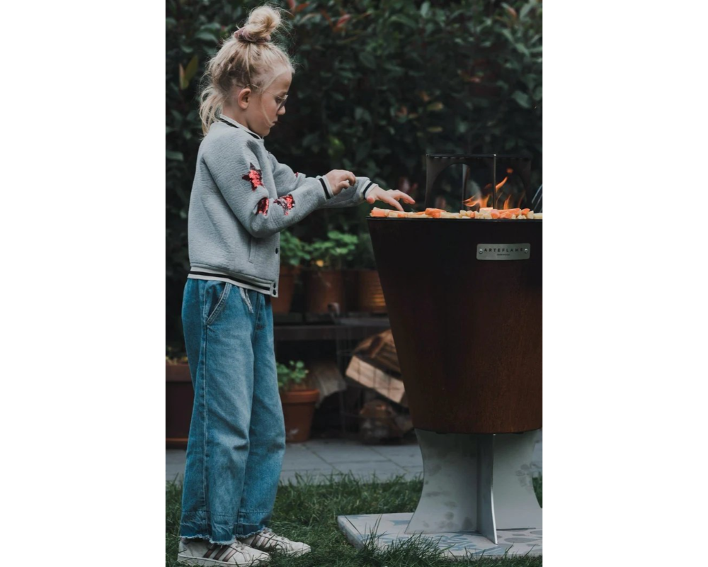 Arteflame One Series 20" Corten Steel Wood Fire Grill with child grilling vegetables on flat-top surface in backyard garden
