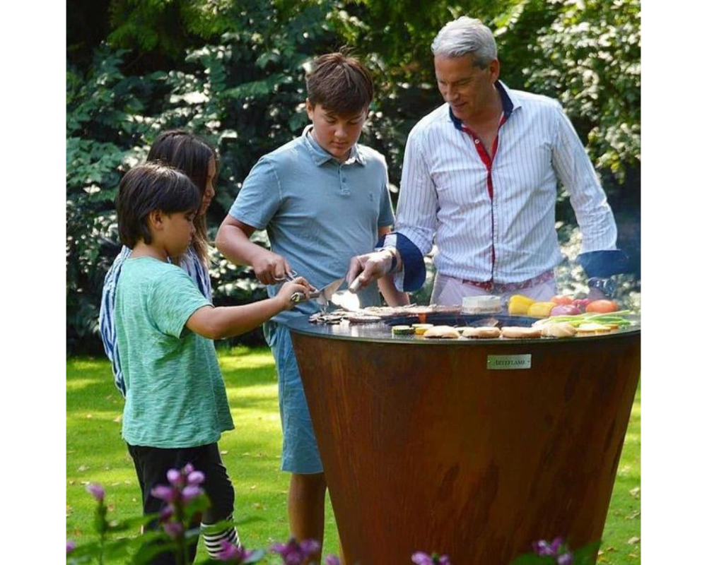 Arteflame One Series 40" Corten Steel Grill with family grilling burgers and vegetables together in backyard garden