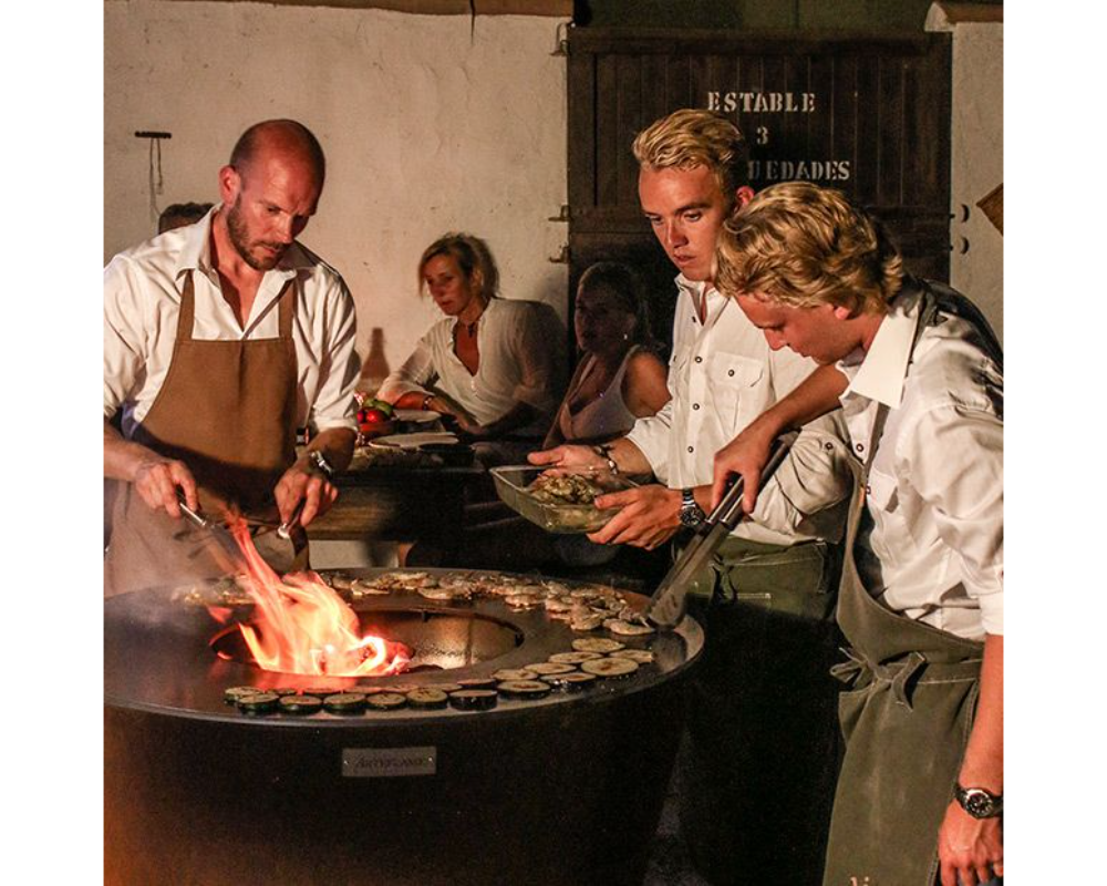 Arteflame One Series 40" Grill with chefs grilling food on flat-top steel surface at outdoor evening social gathering