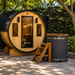 Wood barrel sauna with glass door beside a black cedar cold plunge ice bath in a garden