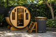 Hot and Cold Bundle: Aleko Barrel Sauna and Cedar Ice Bath, front view of the barrel sauna on a garden patio, black shingled roof, glass door and two windows, potted plants beside it