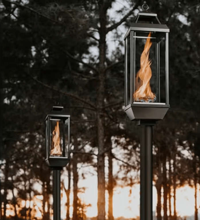 Pair of Travis Industries Tempest gas lanterns on in-ground posts with vortex flames burning at dusk in wooded outdoor setting
