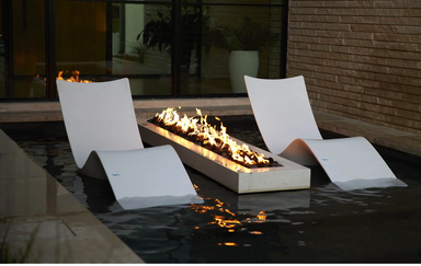 Two Ledge Lounger Autograph Chaises set in shallow water on either side of a linear fire feature