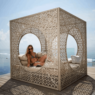A woman in a bikini sits inside a large, cream-colored outdoor cube daybed with intricate laser-cut geometric patterns. The daybed is situated on a wooden deck with the ocean and blue sky visible in the background.