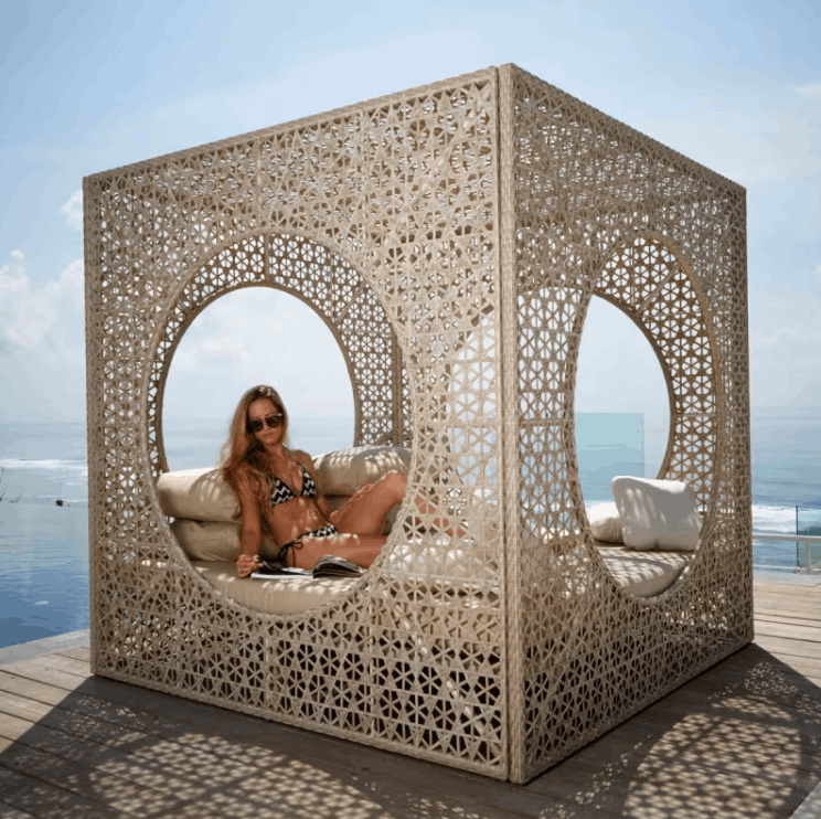 A woman in a bikini sits inside a large, cream-colored outdoor cube daybed with intricate laser-cut geometric patterns. The daybed is situated on a wooden deck with the ocean and blue sky visible in the background.