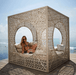 A woman in a bikini sits inside a large, cream-colored outdoor cube daybed with intricate laser-cut geometric patterns. The daybed is situated on a wooden deck with the ocean and blue sky visible in the background.