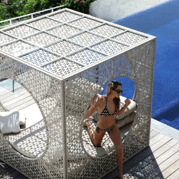 A woman in a black and white patterned bikini sits inside a large, white, geometric outdoor lounge cabana with a laser-cut metal lattice design, positioned next to a blue swimming pool on a wooden deck.