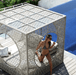 A woman in a black and white patterned bikini sits inside a large, white, geometric outdoor lounge cabana with a laser-cut metal lattice design, positioned next to a blue swimming pool on a wooden deck.