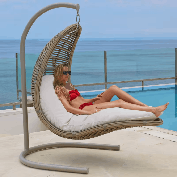 A woman in a red bikini lounges in a woven hanging daybed with white cushions, set on a patio with a pool and ocean in the background.