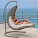 A woman in a red bikini lounges in a woven hanging daybed with white cushions, set on a patio with a pool and ocean in the background.
