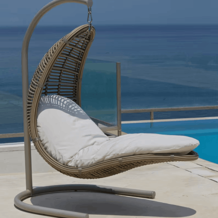 A hanging wicker egg chair with a white cushion, suspended by a curved metal frame on a patio with a blue ocean and pool in the background.