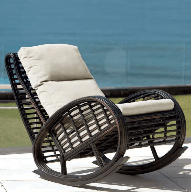 A dark brown wicker rocking chair with cream-colored cushions, positioned outdoors on a patio with a blue ocean background.