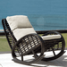 A dark brown wicker rocking chair with cream-colored cushions, positioned outdoors on a patio with a blue ocean background.