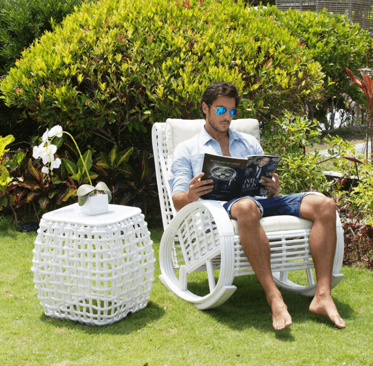 A man wearing blue sunglasses sits barefoot in a white outdoor rocking chair reading a book, with a matching white woven side table beside him on a grassy lawn in front of green bushes.