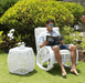 A man wearing blue sunglasses sits barefoot in a white outdoor rocking chair reading a book, with a matching white woven side table beside him on a grassy lawn in front of green bushes.