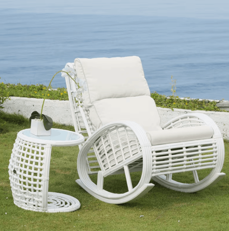 White wicker outdoor rocking chair with matching cushioned seat and back, shown on grass beside a small round side table, with the ocean and low wall in the background.