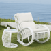 White wicker outdoor rocking chair with matching cushioned seat and back, shown on grass beside a small round side table, with the ocean and low wall in the background.