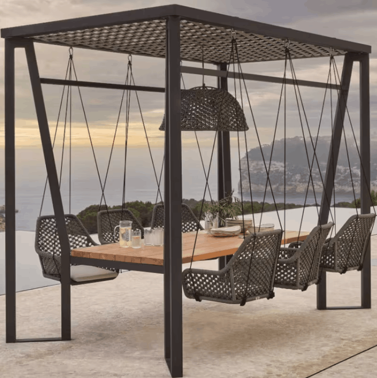A large outdoor swing cabana with a suspended dining table and six hanging woven chairs, set on a patio overlooking a scenic ocean and mountain view.