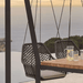 A dark woven outdoor hanging swing chair with a light cushion sits beside a wooden table on a patio, overlooking a sea view at sunset.