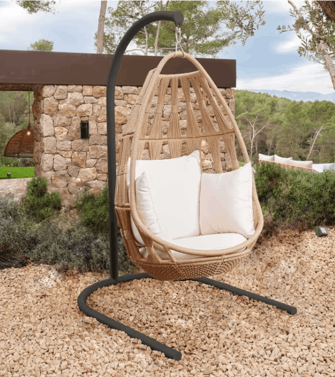 A hanging egg chair with a natural woven rattan body, two white throw pillows, and a black metal curved stand, set outdoors on a gravel surface in front of a stone wall and trees.