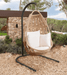 A hanging egg chair with a natural woven rattan body, two white throw pillows, and a black metal curved stand, set outdoors on a gravel surface in front of a stone wall and trees.