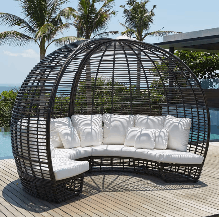 A curved outdoor wicker daybed with a dome-shaped overhead cage canopy, upholstered in white fabric with matching white pillows, set on a wooden deck near a pool with palm trees and ocean in the background.