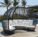 A curved outdoor wicker daybed with a dome-shaped overhead cage canopy, upholstered in white fabric with matching white pillows, set on a wooden deck near a pool with palm trees and ocean in the background.