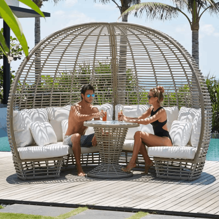 A large dome-shaped outdoor wicker lounge chair with white cushions, shown on a wooden deck by a pool with a man and woman sitting inside and having a drink.