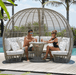A large dome-shaped outdoor wicker lounge chair with white cushions, shown on a wooden deck by a pool with a man and woman sitting inside and having a drink.