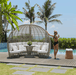 A woman in a black swimsuit stands beside a large oval wicker daybed with white cushions on a wooden pool deck, with palm trees, ocean, and a blue sky in the background.