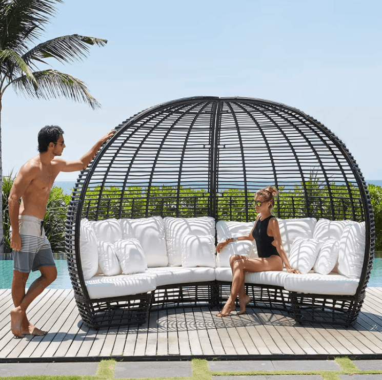 A large black oval wicker lounge chair with white cushions sits on a wooden deck beside a pool, with a man standing on the left and a woman sitting on the right.